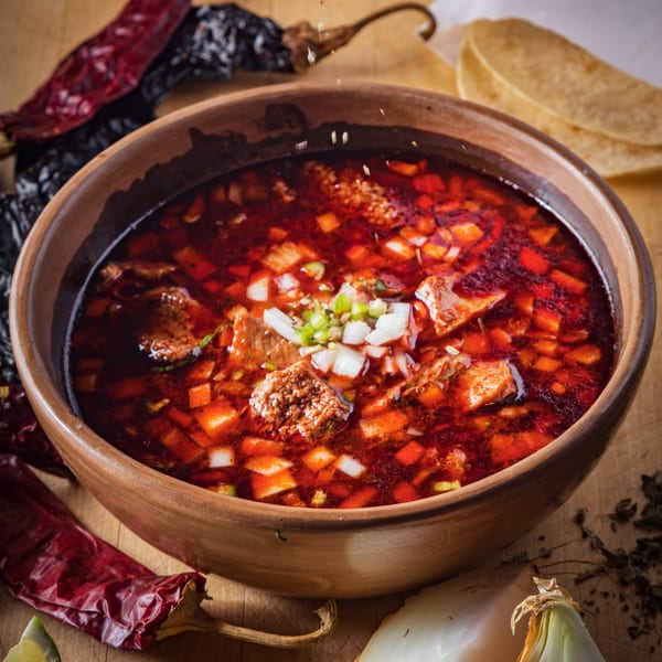 A rustic bowl of red birria stew, garnished with diced onions and cilantro, with tortillas and dried chilies.