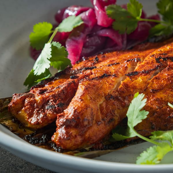 Grilled fish with red seasoning, served on a banana leaf with pickled red onions and cilantro.