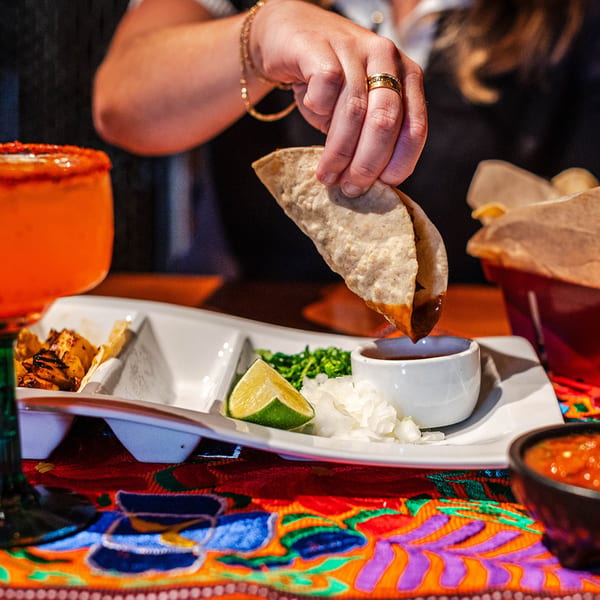A hand dips a Picos Restaurant taco into a small bowl of sauce, with a margarita nearby.